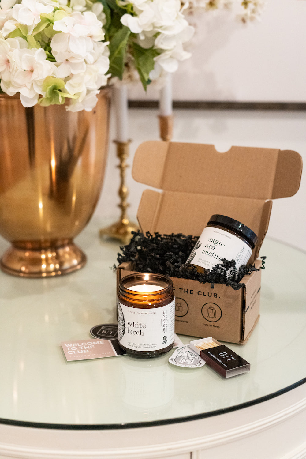 9oz White Birch Soy Candle burning on a glass top next to a matchbook, stickers, and a Candle Club shipping box containing a 9oz Saguaro Cactus Soy Candle.