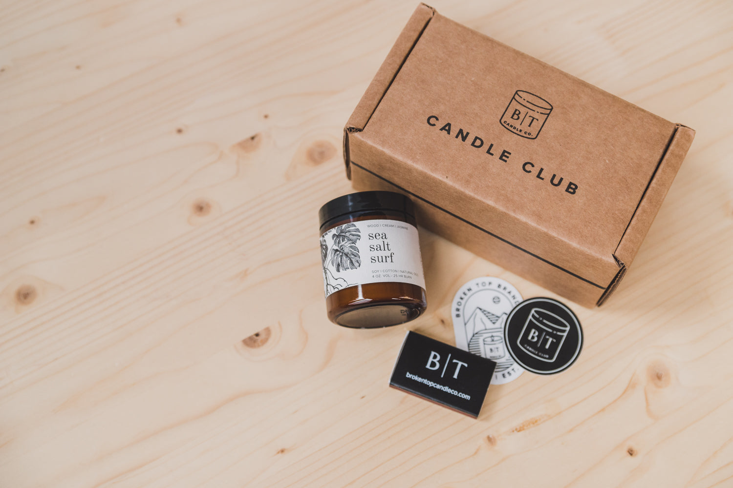 9oz Sea Salt Surf Soy Candle on a wooden table next to stickers, a matchbook, and a Candle Club shipping box. 