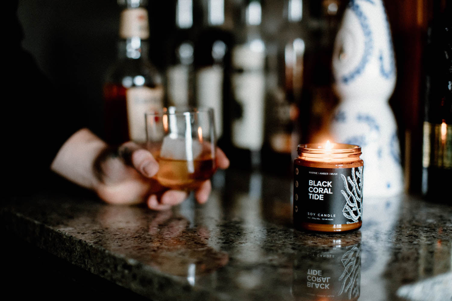 9oz Black Coral Tide Soy Candle on a countertop in front of a whiskey glass being held by a hand. 