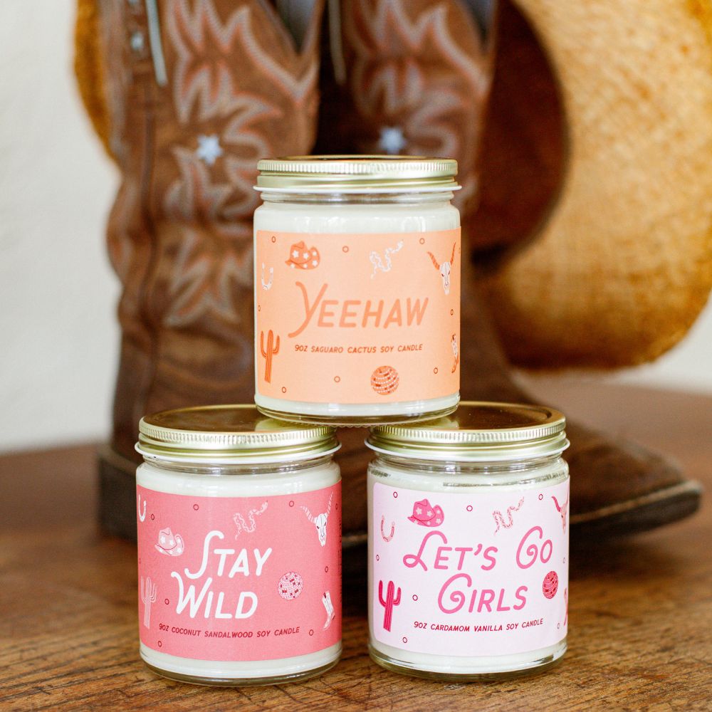 9oz &#39;Yeehaw&#39;, &#39;Stay Wild&#39;, and &#39;Let&#39;s Go Girls&#39; Soy Candles in front of a pair of cowboy boots and hat. 