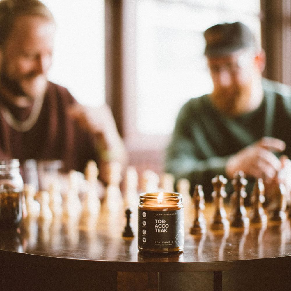 9oz Tobacco Teak Soy Candle in front of two men playing chess. 