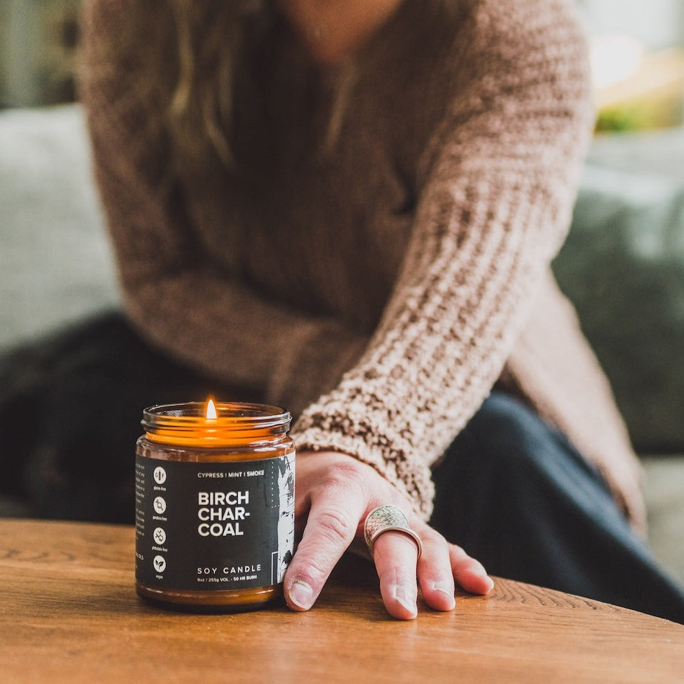 9oz Birch Charcoal Soy Candle on a coffee table being held by a woman's hand. 
