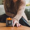 9oz Birch Charcoal Soy Candle on a coffee table being held by a woman's hand. 