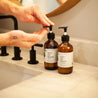 8oz Apricot Bloom Liquid Soap next to a bathroom sink being dispensed by two hands. 