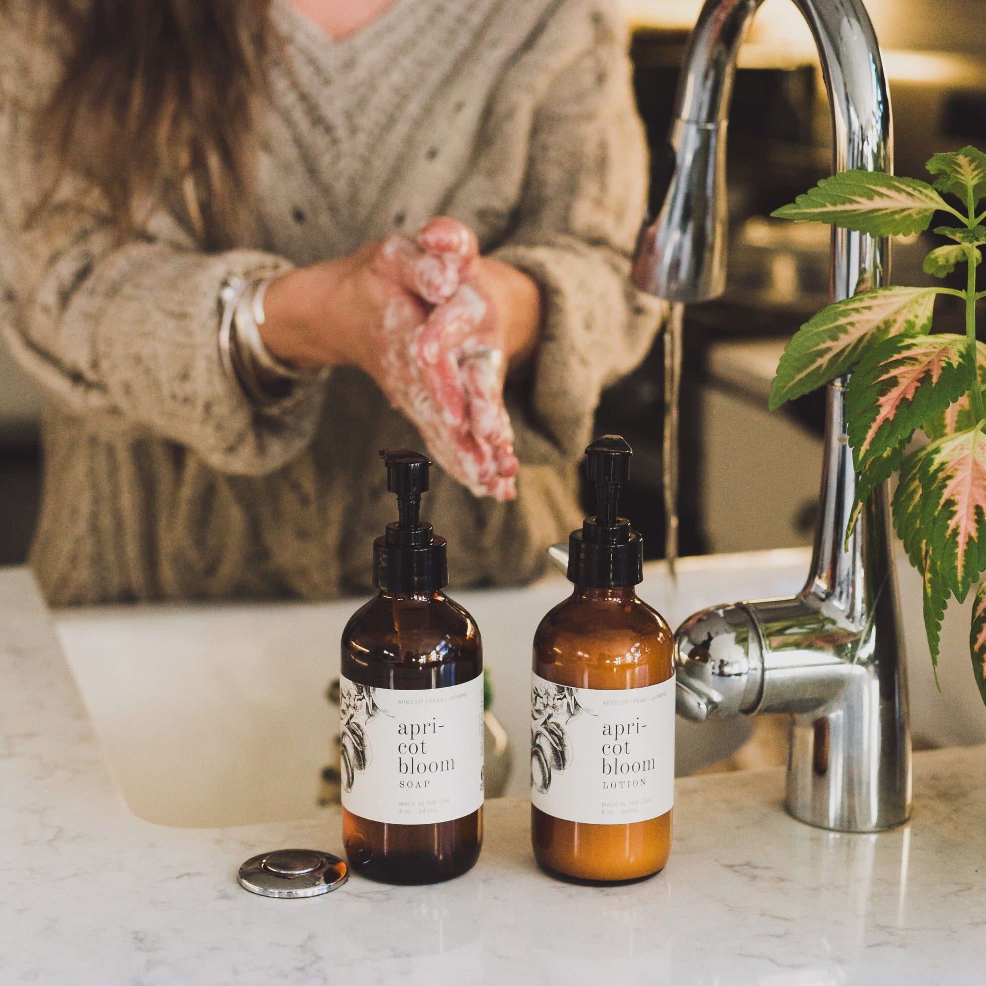 Apricot Bloom 8oz Liquid Soap and 8oz Apricot Bloom Body Lotion in front of a kitchen sink with a woman washing her hands. 