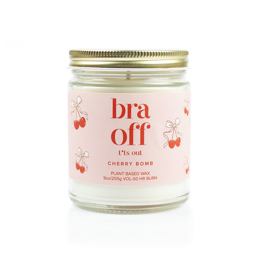 A plant-based wax candle in a clear glass jar with a gold metal lid and a light pink label. The label features the text 'bra off' in red, 't's out' in black, and 'CHERRY BOMB' in red, decorated with cherry illustrations.