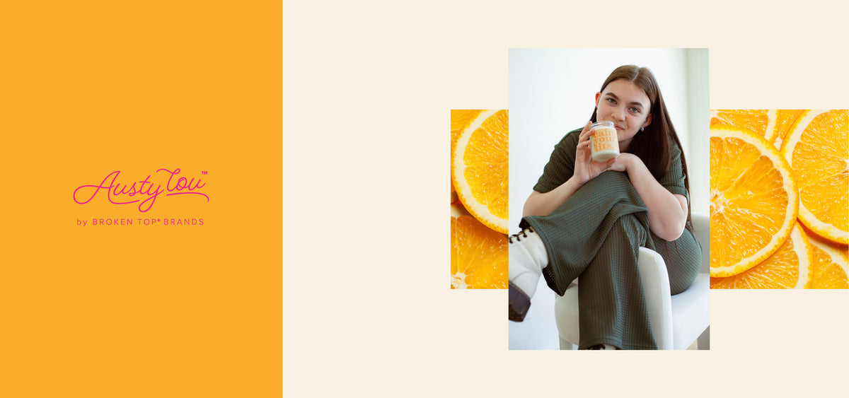 Austy Lou by Broken Top Brands logo. Young woman holding 9oz 'Calm Your Tits' Soy Candle on background of orange slices. 