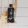 16oz Birch Charcoal Body wash in a shower caddy next to a luffa and washcloth. 