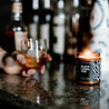 9oz Black Coral Tide Soy Candle on a countertop next to a whisky glass held by a hand. 