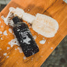 16oz Black Coral Tide Body Wash on wooden bench next to a luffa. 