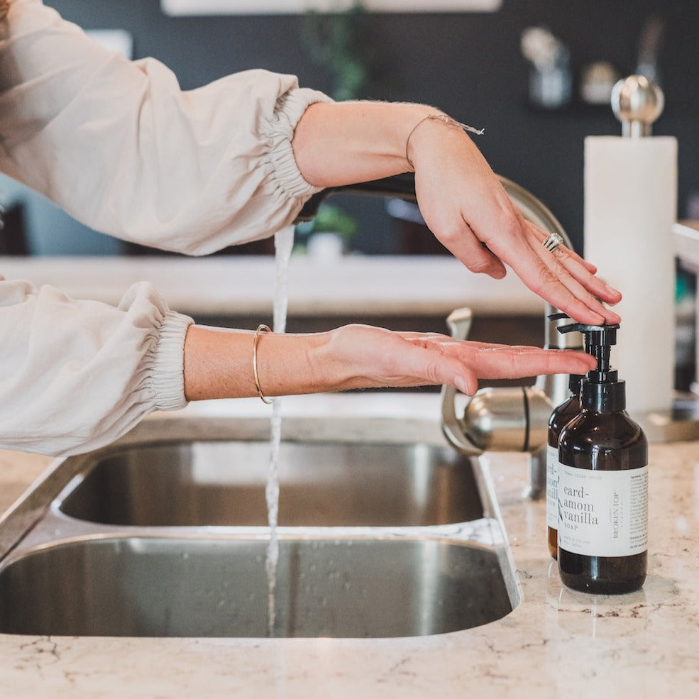 8oz Cardamom Vanilla Liquid Soap next to a kitchen sink being dispensed by two hands. 