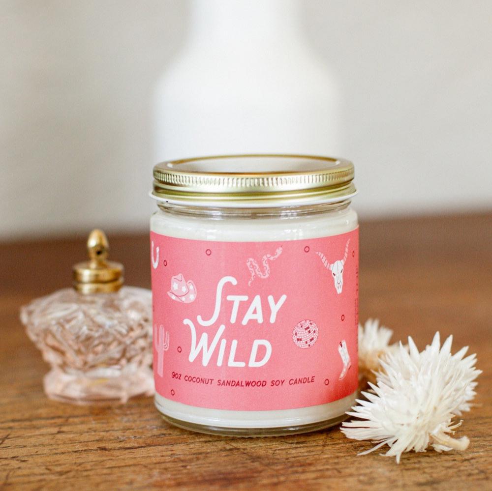 9oz 'Stay Wild' Soy Candle on a wooden table next to decorative glass jar and flowers. 