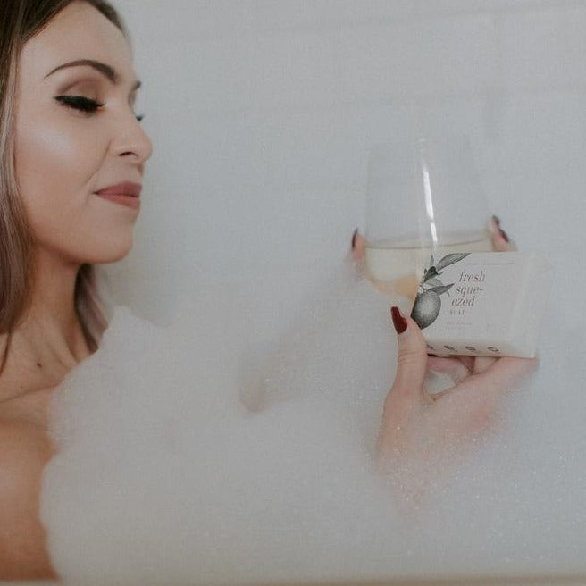 Fresh Squeezed Bar Soap being held by a woman sipping wine in a bathtub. 