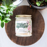 'Tough As A Mother' 9oz Soy Candle on a decorative wooden shelf next to potted plants. 