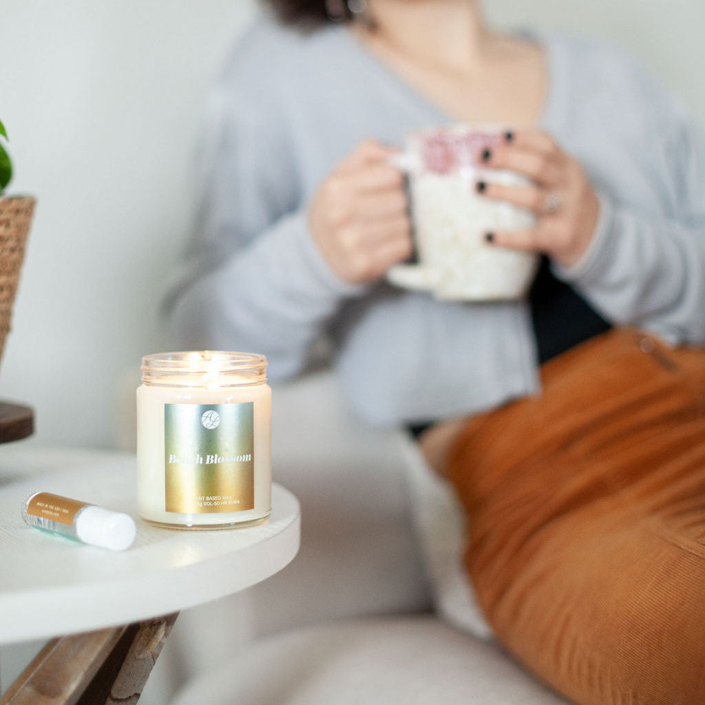 9oz Austy Lou Beach Blossom Soy Candle on a side table next to a Roll On Perfume in front of a young woman drinking from a mug. 