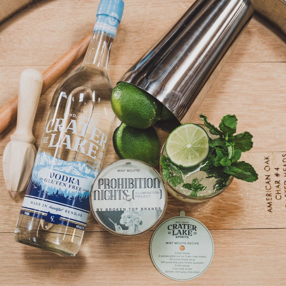11oz Mint Mojito Soy Candle on a barrel with Crater Lake Vodka, cocktail shaker, limes, mint, and a Mint Mojito cocktail. 