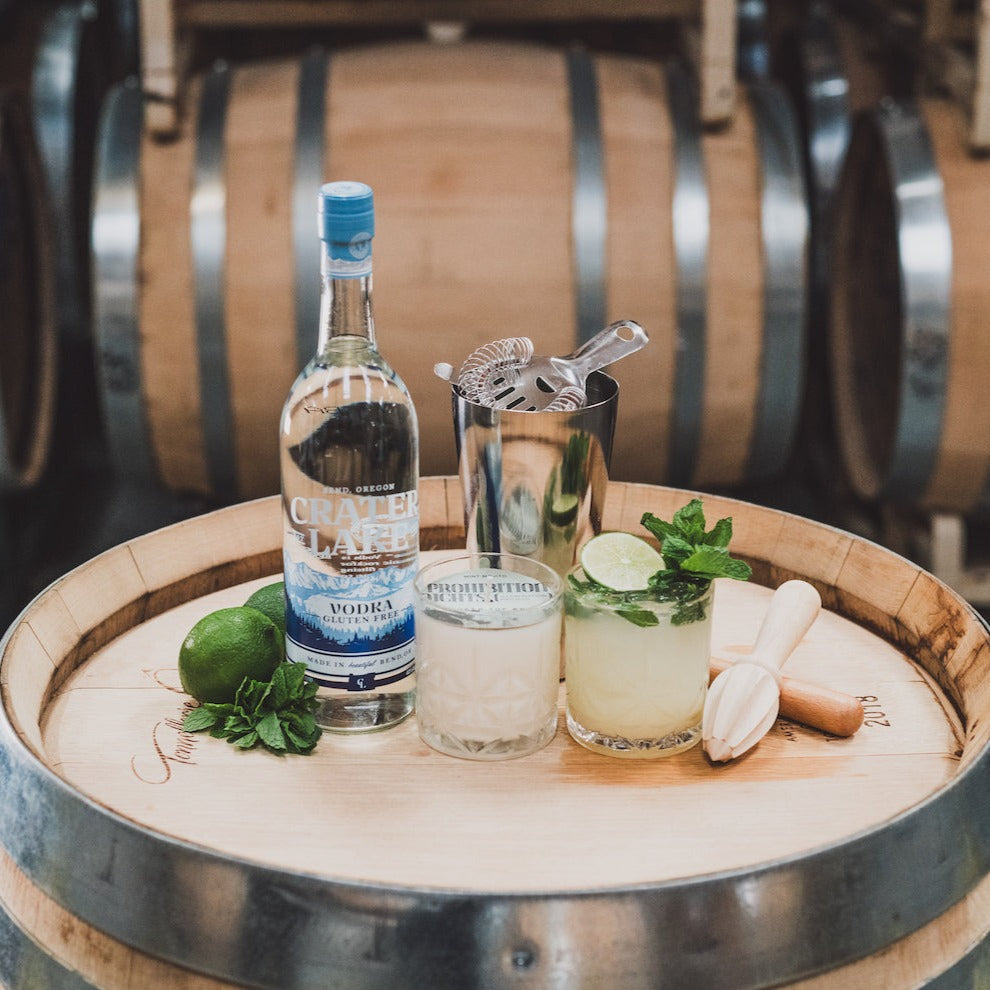 11oz Mint Mojito Soy Candle on a barrel with Crater Lake Vodka, cocktail shaker, limes, mint, and a Mint Mojito cocktail. 
