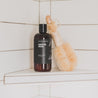 16oz Santal Noir Body Wash next to a luffa in a white tiled shower. 