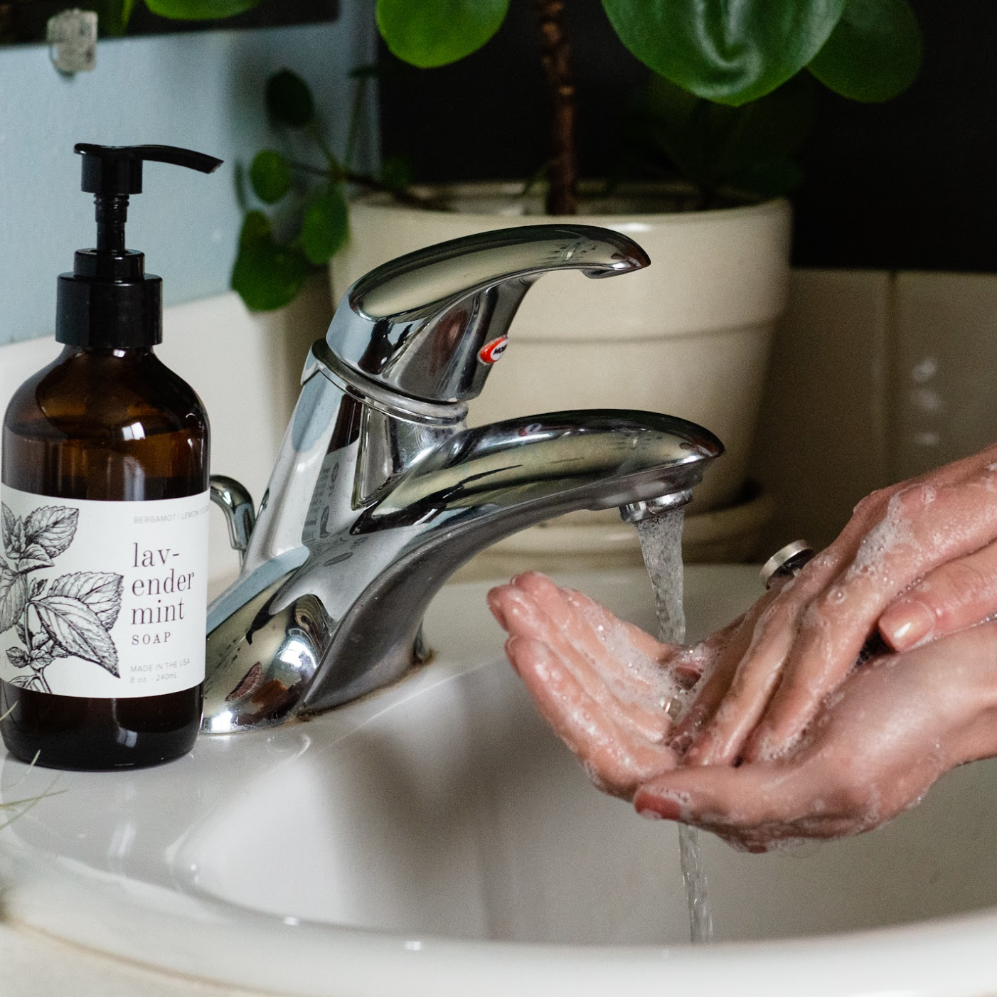 8oz Lavender Mint Liquid Soap next to a bathroom sink with hands washing. 