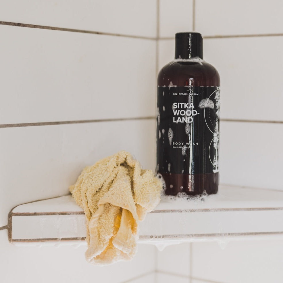 16oz Sitka Woodland Body Wash in white tiled shower next to a washcloth. 