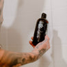 16oz Sitka Woodland Body Wash being held by a man in the shower. 