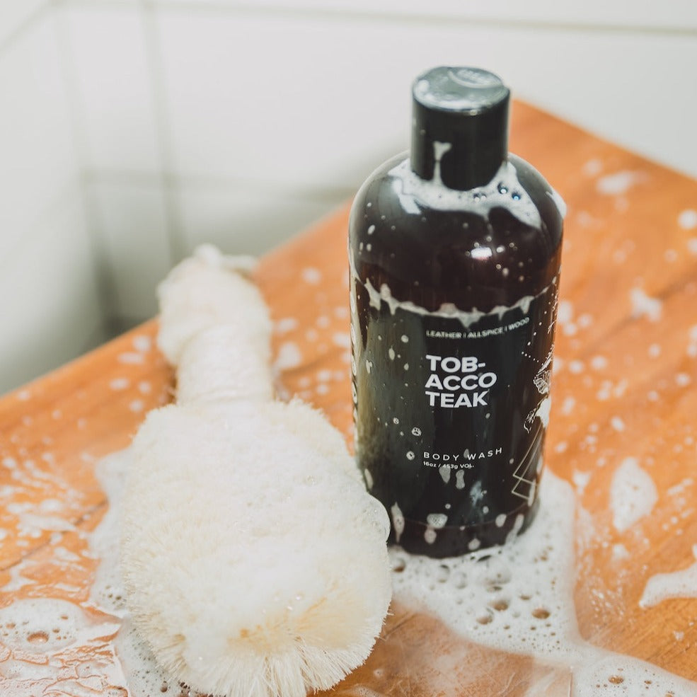16oz Tobacco Teak Body Wash in a shower next to a soapy luffa.