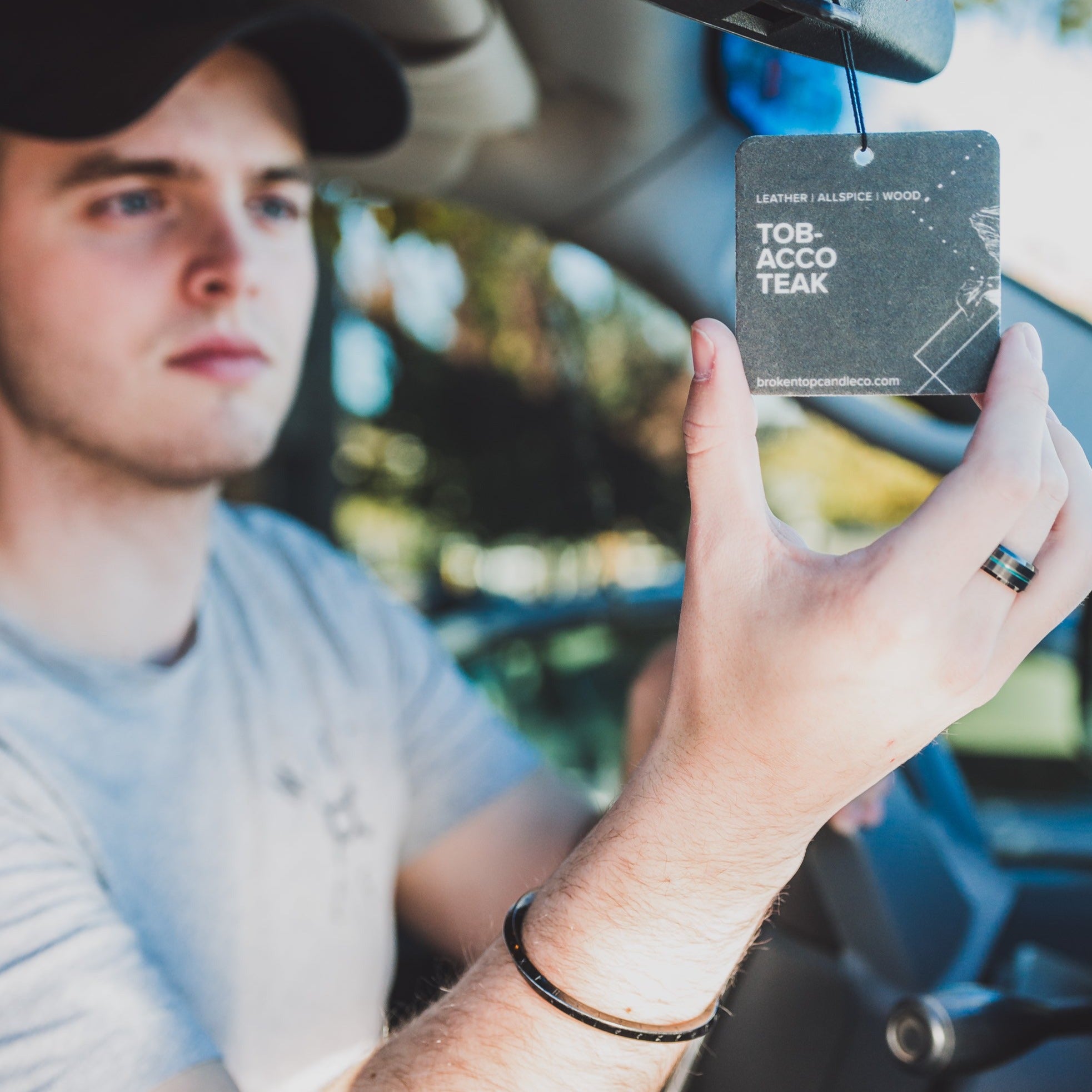Tobacco Teak Car Freshener being hung on a rearview mirror by a man.